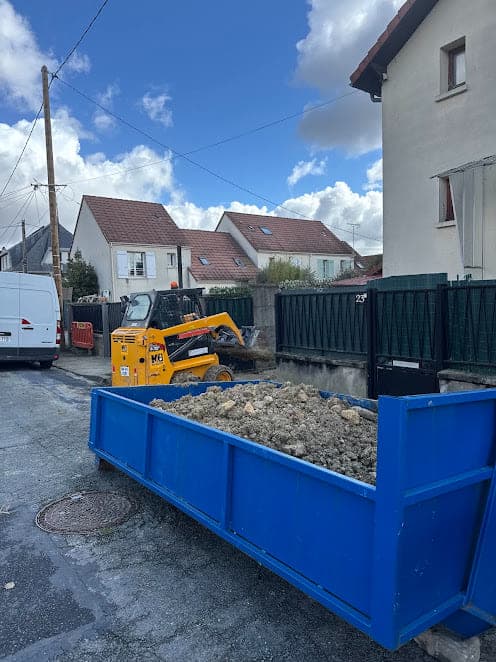 Benne bleue remplie de gravats dans une rue pavillonnaire, chargeuse compacte jaune à l'arrière-plan