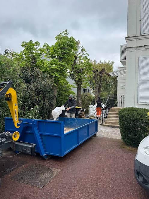 Benne bleue posée par un bras de grue jaune dans l'allée d'une propriété, deux compagnons chargent des sacs de gravats