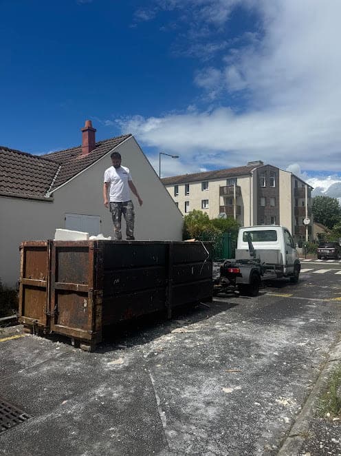 Compagnon debout sur une benne noire pleine de gravats devant un pavillon, camion ampliroll blanc stationné à l'arrière